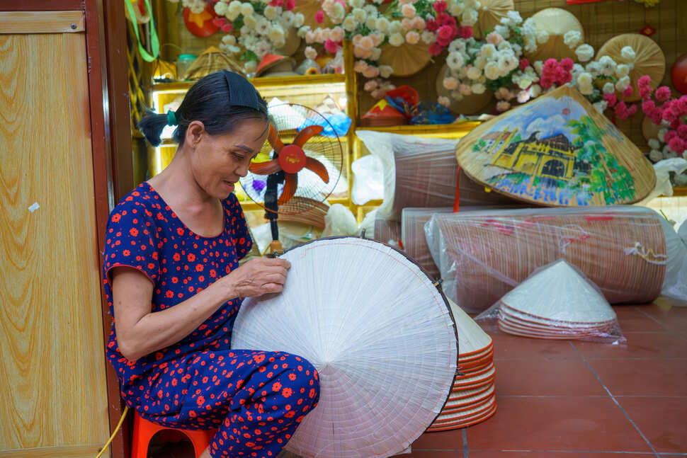 Phía ngoài cổng chợ, không gian trưng bày trở thành điểm dừng chân thu hút du khách. Tại đây, nghệ nhân trực tiếp trình diễn kỹ thuật thêu nón thủ công, đồng thời hướng dẫn khách tham quan trải nghiệm quy trình làm nón và lựa chọn những sản phẩm làm quà lưu niệm. Ảnh: Nguyễn Thuý
